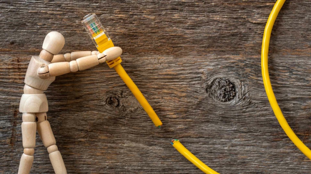 Wooden mannequin uses an electrical tester on a yellow cable over rustic wood, suggesting DIY repair, electrical work and caution.