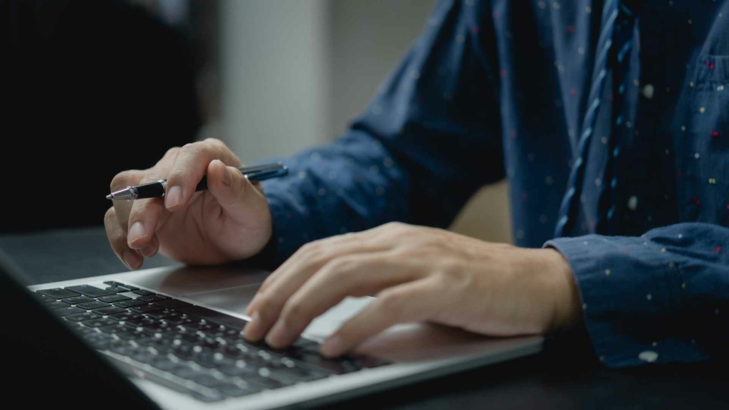A person wearing a blue shirt types on a laptop keyboard and holds a pen in their right hand, possibly researching the upcoming copper broadband switch-off.