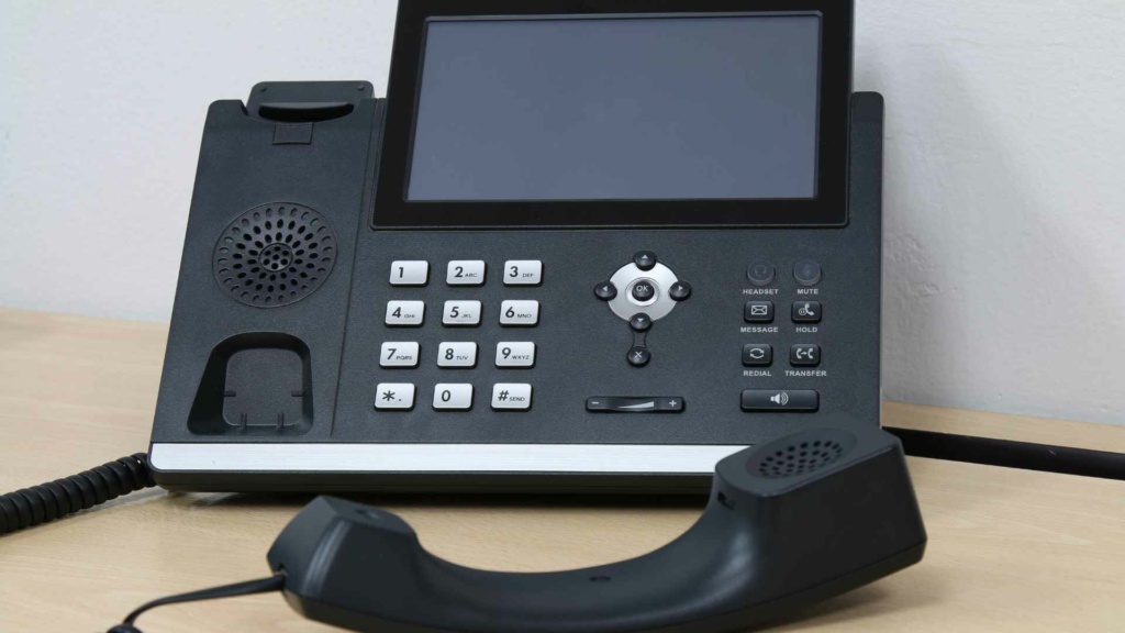 A black office desk VoIP phone with a digital screen and detached handset resting on a wooden surface.