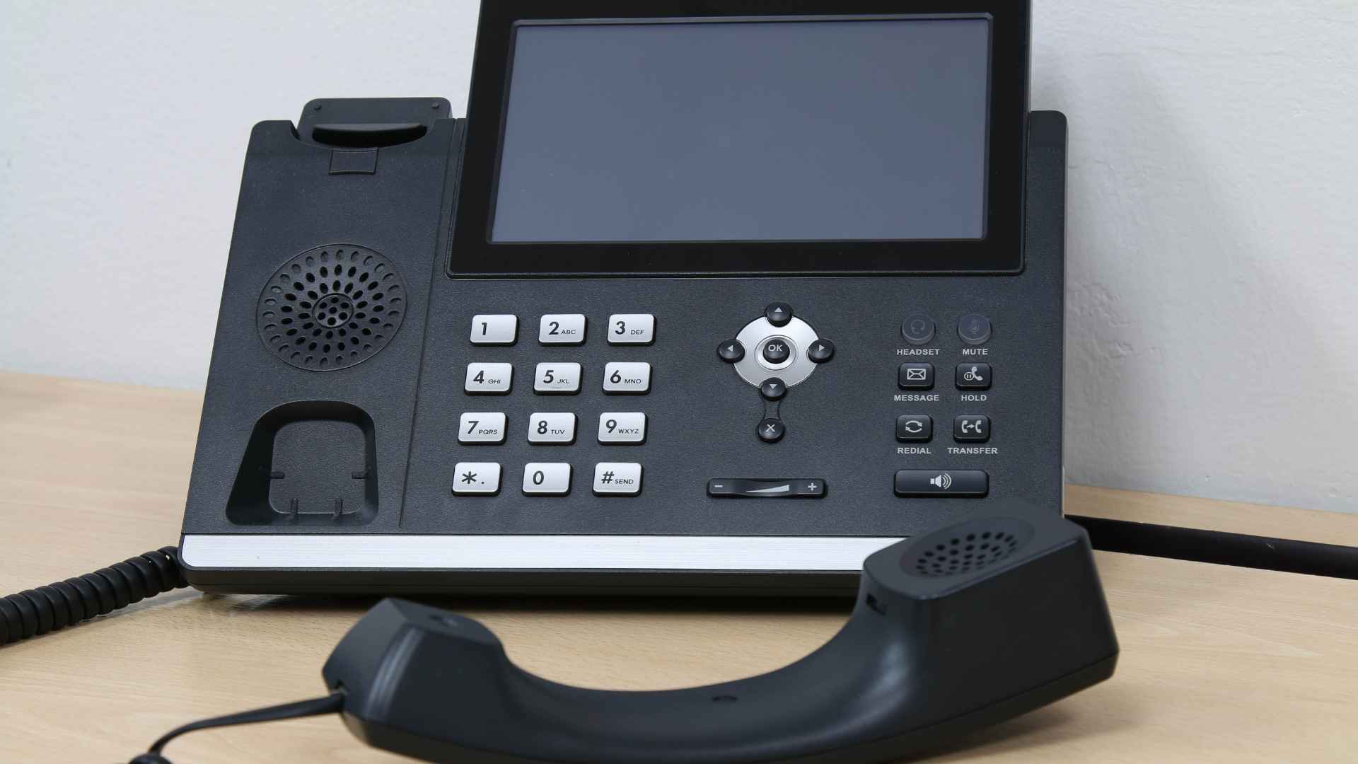 A black office desk VoIP phone with a digital screen and detached handset resting on a wooden surface.
