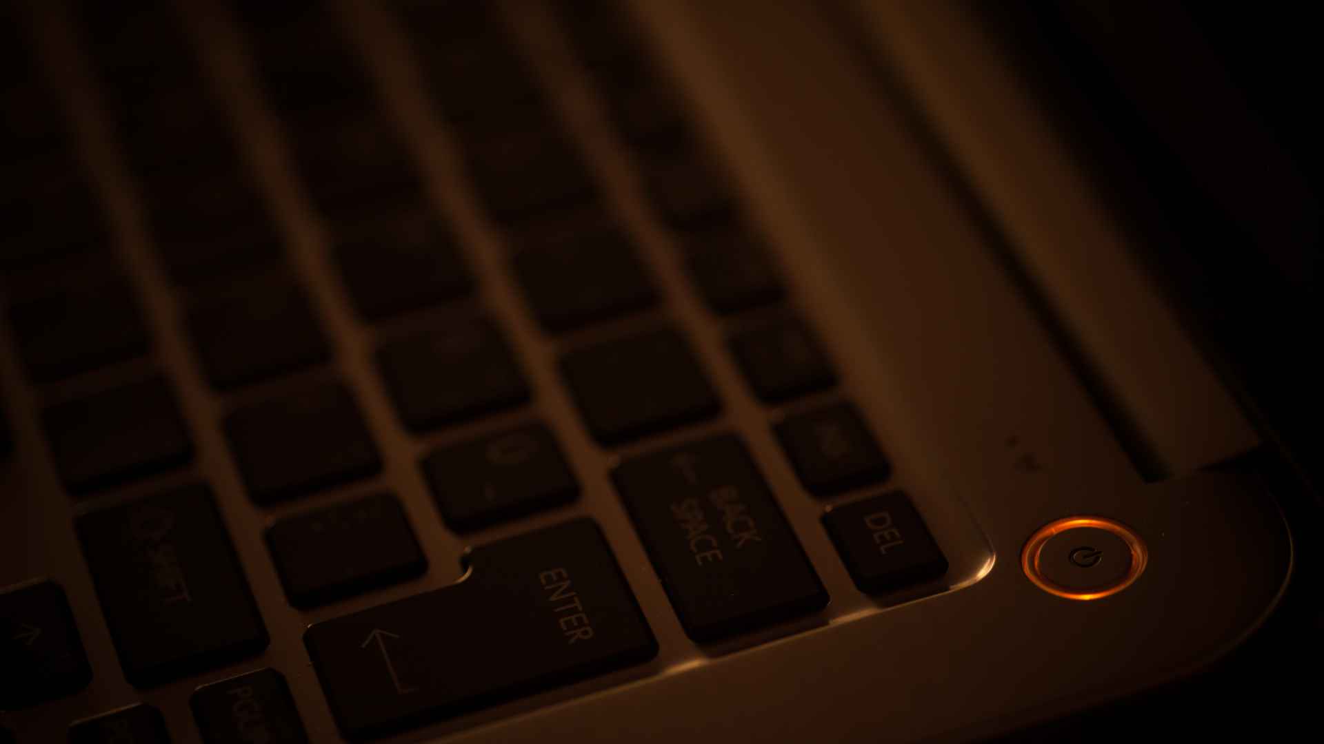 Close-up of a laptop keyboard in dim, warm lighting, with an orange-glowing power button creating a sleek, moody atmosphere.