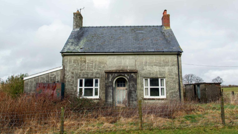 Weathered rural cottage office with stone walls, slate roof, arched doorway, white-framed windows, overgrown yard, and cloudy gray sky.