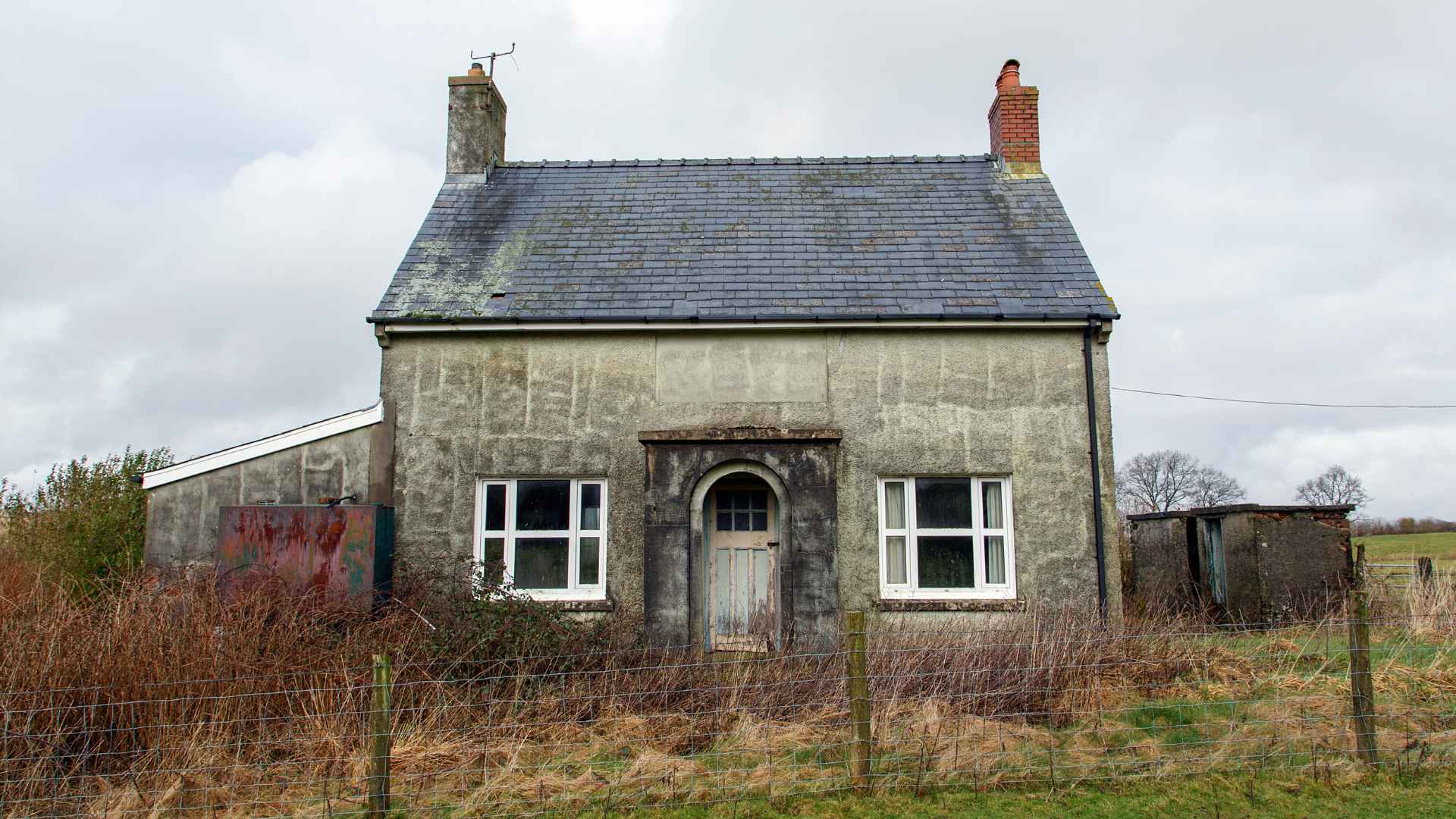 Weathered rural cottage office with stone walls, slate roof, arched doorway, white-framed windows, overgrown yard, and cloudy gray sky.